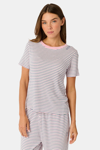 A person stands barefoot on a white background wearing the Roller Rabbit Striped Eva Pant—loose, light-colored pima cotton pajama pants featuring thin blue and red horizontal stripes. Their top is lifted slightly, revealing the elastic waistband.
