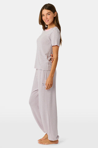 A person stands barefoot on a white background wearing the Roller Rabbit Striped Eva Pant—loose, light-colored pima cotton pajama pants featuring thin blue and red horizontal stripes. Their top is lifted slightly, revealing the elastic waistband.