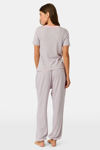 A person stands barefoot on a white background wearing the Roller Rabbit Striped Eva Pant—loose, light-colored pima cotton pajama pants featuring thin blue and red horizontal stripes. Their top is lifted slightly, revealing the elastic waistband.