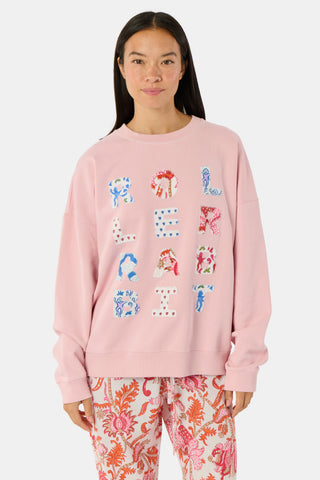 A woman stands against a white background wearing the Roller Rabbit Applique Crewneck by Roller Rabbit—a light pink sweater featuring colorful, patterned letters spelling “ROLLERRABBIT”—paired with pink and orange floral pants.