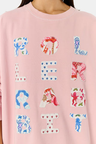 A woman stands against a white background wearing the Roller Rabbit Applique Crewneck by Roller Rabbit—a light pink sweater featuring colorful, patterned letters spelling “ROLLERRABBIT”—paired with pink and orange floral pants.