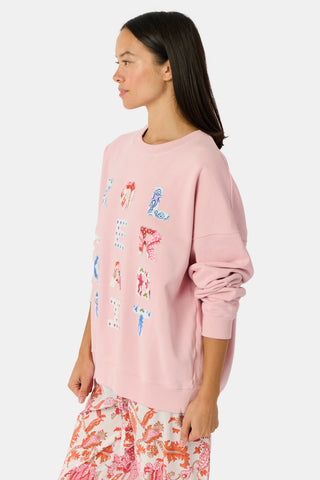 A woman stands against a white background wearing the Roller Rabbit Applique Crewneck by Roller Rabbit—a light pink sweater featuring colorful, patterned letters spelling “ROLLERRABBIT”—paired with pink and orange floral pants.