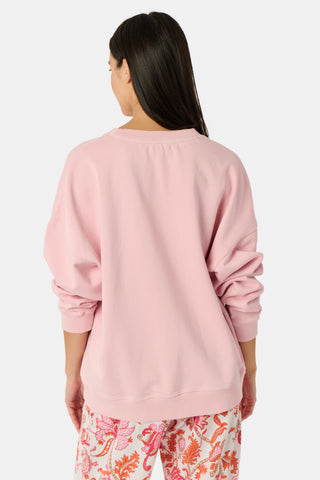A woman stands against a white background wearing the Roller Rabbit Applique Crewneck by Roller Rabbit—a light pink sweater featuring colorful, patterned letters spelling “ROLLERRABBIT”—paired with pink and orange floral pants.