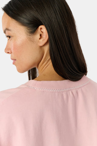 A woman stands against a white background wearing the Roller Rabbit Applique Crewneck by Roller Rabbit—a light pink sweater featuring colorful, patterned letters spelling “ROLLERRABBIT”—paired with pink and orange floral pants.