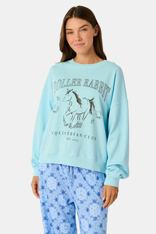 Wearing the Roller Rabbit Equestrian Club Phoebe Sweatshirt in baby blue, a woman pairs it with horse-patterned shorts as she holds a baby goat. She stands by a wooden fence with greenery in the background, smiling softly.