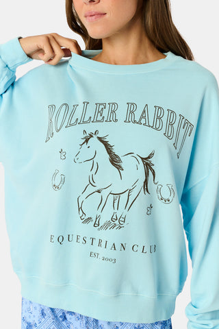Wearing the Roller Rabbit Equestrian Club Phoebe Sweatshirt in baby blue, a woman pairs it with horse-patterned shorts as she holds a baby goat. She stands by a wooden fence with greenery in the background, smiling softly.