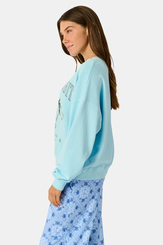 Wearing the Roller Rabbit Equestrian Club Phoebe Sweatshirt in baby blue, a woman pairs it with horse-patterned shorts as she holds a baby goat. She stands by a wooden fence with greenery in the background, smiling softly.
