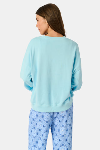 Wearing the Roller Rabbit Equestrian Club Phoebe Sweatshirt in baby blue, a woman pairs it with horse-patterned shorts as she holds a baby goat. She stands by a wooden fence with greenery in the background, smiling softly.
