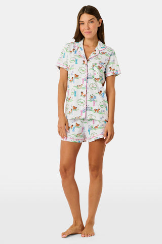 Introducing the Saddle Up Polo Pajamas by Roller Rabbit: a short-sleeve, button-up set with coordinating shorts. Crafted from white fabric adorned with a vibrant equestrian print—showcasing colorful horses, trees, and fences—these pajamas are finished with playful pink trim along the edges and collar. Perfect for horse-girl PJ fans and lovers of both Western and English riding styles.