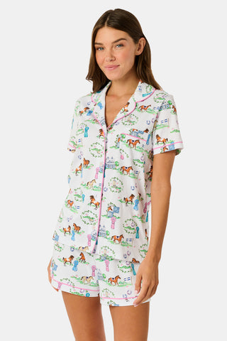 Introducing the Saddle Up Polo Pajamas by Roller Rabbit: a short-sleeve, button-up set with coordinating shorts. Crafted from white fabric adorned with a vibrant equestrian print—showcasing colorful horses, trees, and fences—these pajamas are finished with playful pink trim along the edges and collar. Perfect for horse-girl PJ fans and lovers of both Western and English riding styles.