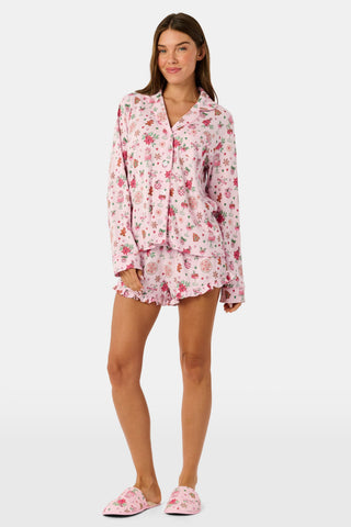 Two women sit on a patterned blanket outdoors wearing matching light pink Seasons Soiree Hayden Pajamas by NETTALCO, featuring a classic Pima cotton top and ruffled shorts decorated with holiday-themed illustrations.