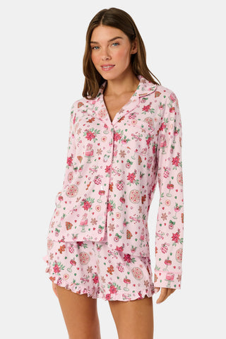 Two women sit on a patterned blanket outdoors wearing matching light pink Seasons Soiree Hayden Pajamas by NETTALCO, featuring a classic Pima cotton top and ruffled shorts decorated with holiday-themed illustrations.