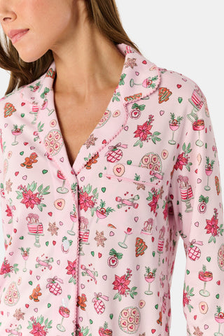 Two women sit on a patterned blanket outdoors wearing matching light pink Seasons Soiree Hayden Pajamas by NETTALCO, featuring a classic Pima cotton top and ruffled shorts decorated with holiday-themed illustrations.
