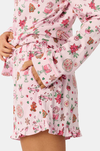 Two women sit on a patterned blanket outdoors wearing matching light pink Seasons Soiree Hayden Pajamas by NETTALCO, featuring a classic Pima cotton top and ruffled shorts decorated with holiday-themed illustrations.