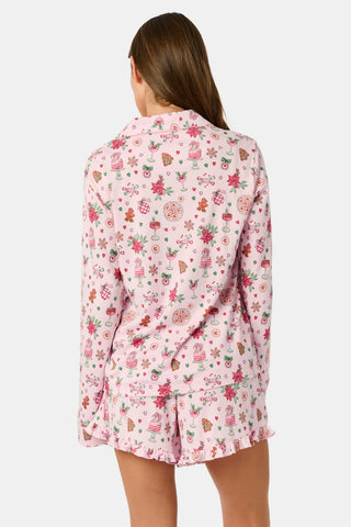 Two women sit on a patterned blanket outdoors wearing matching light pink Seasons Soiree Hayden Pajamas by NETTALCO, featuring a classic Pima cotton top and ruffled shorts decorated with holiday-themed illustrations.