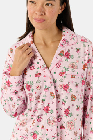 A person wearing pink long-sleeved Seasons Soiree Kate Pajamas with a ruffled hem and a repeating print of holiday sweets and treats.