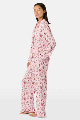 A person wearing pink long-sleeved Seasons Soiree Kate Pajamas with a ruffled hem and a repeating print of holiday sweets and treats.