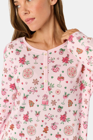 A person with long blonde hair sits on a patterned quilt outdoors, wearing a long-sleeved NETTALCO Seasons Soiree Suzie Sleep Dress featuring small food and drink icons.