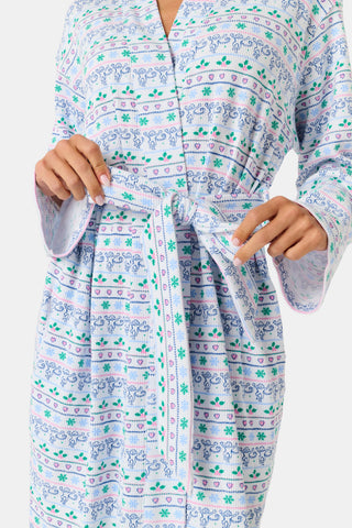 A woman wears a knee-length SERFLEX Snowday Stripe Piper Robe featuring horizontal rows of winter-themed snowflakes, hearts, and stars in shades of blue, pink, and green.