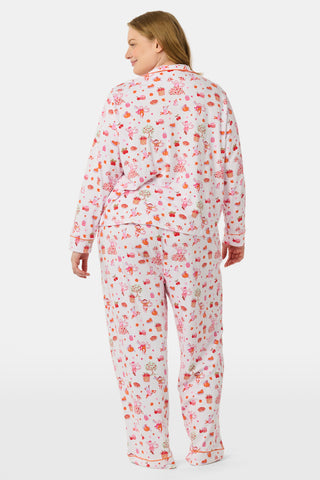 A woman stands barefoot against a plain white background, hands in pockets, wearing the Roller Rabbit Sweater Weather Long Sleeve Polo Pajamas—a floral pink and white set featuring a long-sleeved polo top and matching pants.