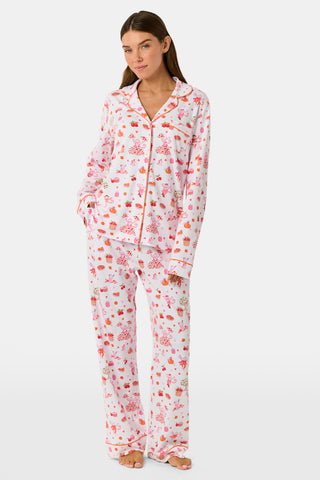 A woman stands barefoot against a plain white background, hands in pockets, wearing the Roller Rabbit Sweater Weather Long Sleeve Polo Pajamas—a floral pink and white set featuring a long-sleeved polo top and matching pants.