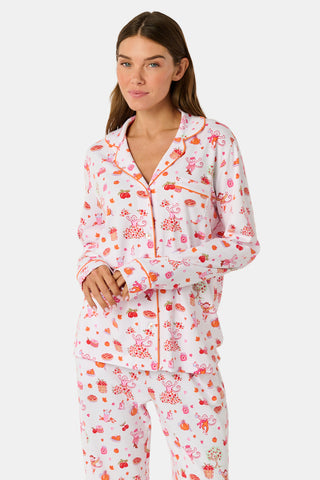 A woman stands barefoot against a plain white background, hands in pockets, wearing the Roller Rabbit Sweater Weather Long Sleeve Polo Pajamas—a floral pink and white set featuring a long-sleeved polo top and matching pants.