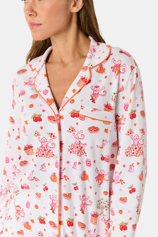 A woman stands barefoot against a plain white background, hands in pockets, wearing the Roller Rabbit Sweater Weather Long Sleeve Polo Pajamas—a floral pink and white set featuring a long-sleeved polo top and matching pants.