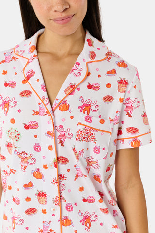 A woman smiles while wearing the Roller Rabbit Sweater Weather Polo Pajamas, a matching set of printed Pima PJs featuring colorful pink and orange cupcakes, flowers, and playful designs on a white background.