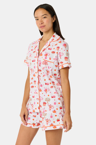 A woman smiles while wearing the Roller Rabbit Sweater Weather Polo Pajamas, a matching set of printed Pima PJs featuring colorful pink and orange cupcakes, flowers, and playful designs on a white background.