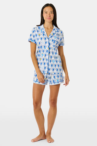 A woman wearing blue and white patterned Tri Delta Monkey Polo Pajamas by Roller Rabbit holds a large striped bag. She stands outdoors near a brick wall and green plants, with sunlight filtering through the leaves.