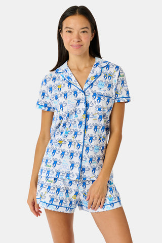 A woman wearing blue and white patterned Tri Delta Monkey Polo Pajamas by Roller Rabbit holds a large striped bag. She stands outdoors near a brick wall and green plants, with sunlight filtering through the leaves.