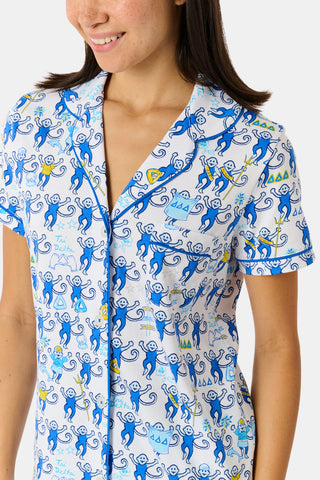 A woman wearing blue and white patterned Tri Delta Monkey Polo Pajamas by Roller Rabbit holds a large striped bag. She stands outdoors near a brick wall and green plants, with sunlight filtering through the leaves.
