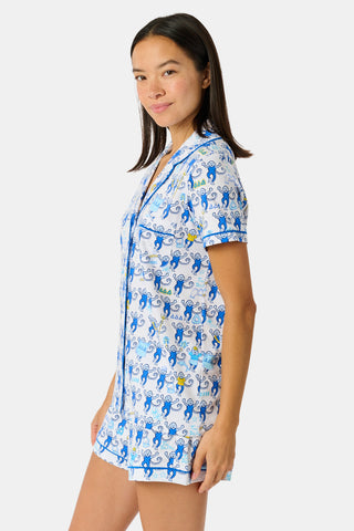 A woman wearing blue and white patterned Tri Delta Monkey Polo Pajamas by Roller Rabbit holds a large striped bag. She stands outdoors near a brick wall and green plants, with sunlight filtering through the leaves.