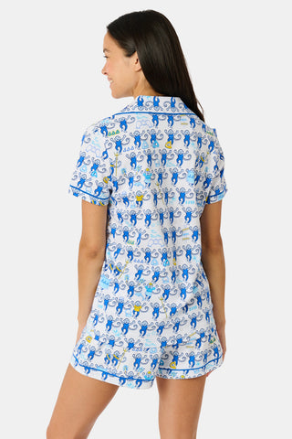 A woman wearing blue and white patterned Tri Delta Monkey Polo Pajamas by Roller Rabbit holds a large striped bag. She stands outdoors near a brick wall and green plants, with sunlight filtering through the leaves.
