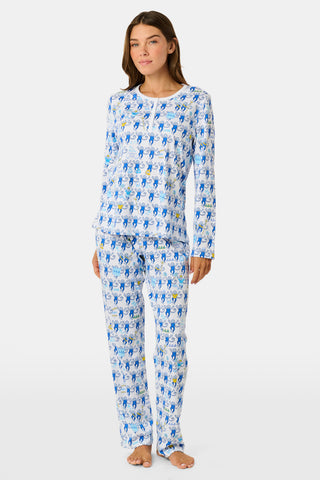 A woman with long brown hair sits on outdoor steps, gazing to the side while effortlessly showcasing her stylish look in Roller Rabbit’s Tri Delta Monkey Pajamas, paired with sunglasses and a navy jacket draped over her shoulders.