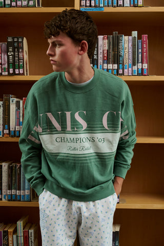 A person wearing a preppy green Roller Rabbit Tennis Champions Andie Sweatshirt stands in front of bookshelves. Only the lower part of their face is visible, and their hands are in their pockets.