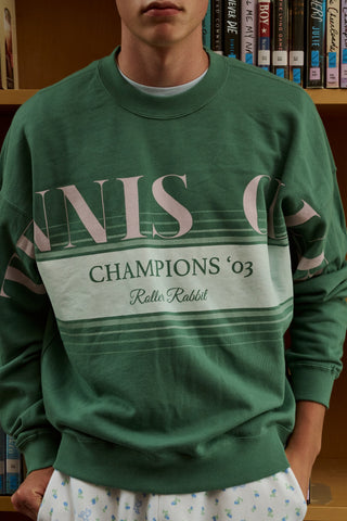 A person wearing a preppy green Roller Rabbit Tennis Champions Andie Sweatshirt stands in front of bookshelves. Only the lower part of their face is visible, and their hands are in their pockets.