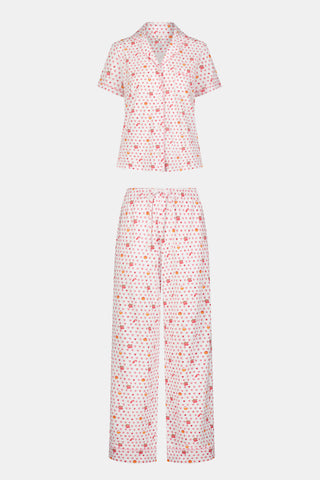 A woman in the Roller Rabbit Autumn Hearts Fiora Pajamas and brown boots sits on a wooden fence, smiling and resting one arm on a tree branch in a sunny outdoor setting, evoking the coziness of fall sleepwear.