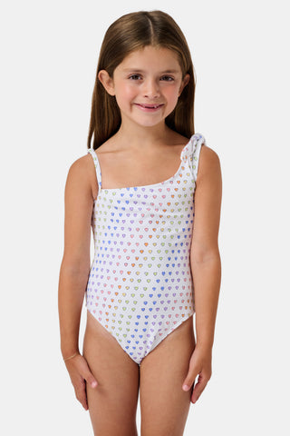 The Disco Hearts Kids Jacey One Piece by Roller Rabbit is a white swimsuit with rows of colorful hearts, featuring an asymmetrical neckline, one thin strap, and a knot-tie detail on the right shoulder.