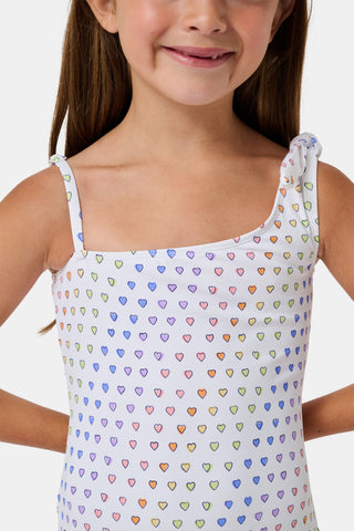 The Disco Hearts Kids Jacey One Piece by Roller Rabbit is a white swimsuit with rows of colorful hearts, featuring an asymmetrical neckline, one thin strap, and a knot-tie detail on the right shoulder.