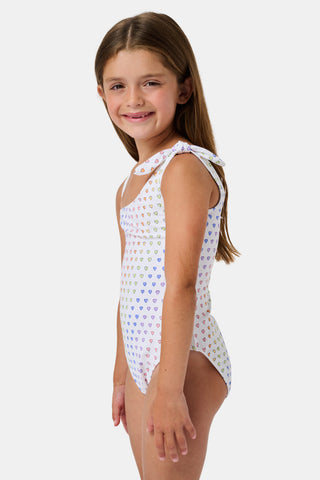 The Disco Hearts Kids Jacey One Piece by Roller Rabbit is a white swimsuit with rows of colorful hearts, featuring an asymmetrical neckline, one thin strap, and a knot-tie detail on the right shoulder.