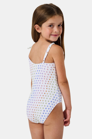 The Disco Hearts Kids Jacey One Piece by Roller Rabbit is a white swimsuit with rows of colorful hearts, featuring an asymmetrical neckline, one thin strap, and a knot-tie detail on the right shoulder.