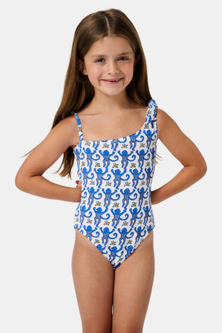 The Monkey Kids Jacey One Piece by Roller Rabbit is a white, one-shoulder swimsuit with blue monkeys and yellow bananas. It features an asymmetrical neckline, a knotted right tie strap, and playful monkey illustrations.
