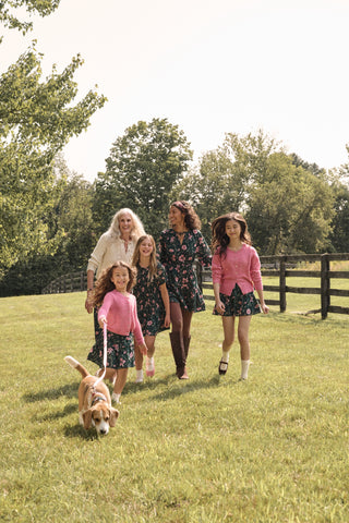 A model wears the SOPHISTICA Greta Floral Belle Dress, a dark green cotton mini dress featuring a pink floral print, a gathered waist, and a ruffled hem.