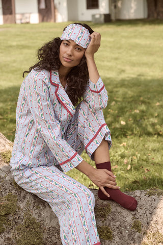 A person wearing a Mistletoe Stripe Josie Loungewear set with red trim, a matching sleep mask, and dark socks, sitting on a mossy tree trunk.