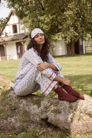 A person wearing a Mistletoe Stripe Josie Loungewear set with red trim, a matching sleep mask, and dark socks, sitting on a mossy tree trunk.