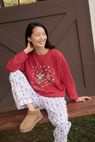 A smiling woman sits on a wooden bench wearing a red Deck The Halls Andie Sweatshirt featuring an illustrated monkey in string lights and patterned Bea Sweatpants.