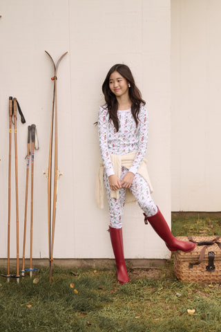 A child wearing SERFLEX Kids Deck The Halls Pajamas, featuring a repeating holiday pattern of snowmen, Christmas trees, and elves connected by garland on long-sleeved Pima cotton.