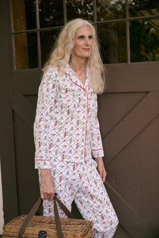 A woman wearing SERFLEX Deck The Halls Long Sleeve Polo Pajamas, a two-piece Pima cotton set featuring a repeating Christmas print of snowmen, elves, and trees on a white background with red piping.