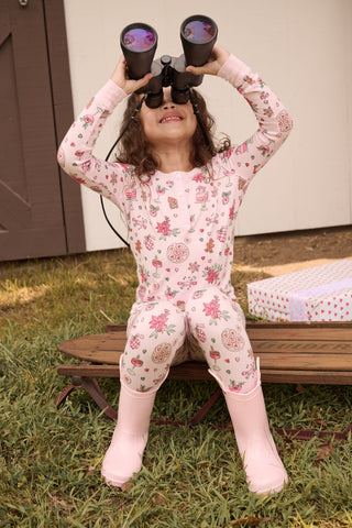 A child in a patterned Kids Seasons Soiree Juniper Pajamas two-piece set and rubber boots sits on a wooden sled and looks through binoculars.